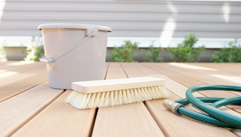 Natural wood deck surface with a soft deck brush, plain bucket, and garden hose resting nearby.