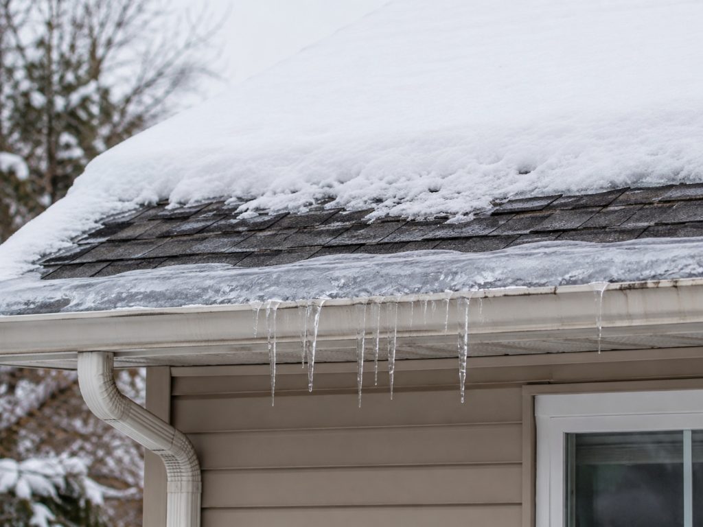 Snow-covered asphalt-shingle roof with ice buildup and icicles forming along the gutter edge.