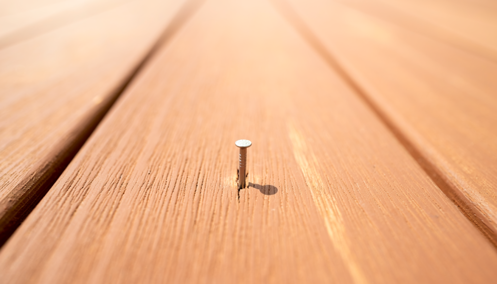 Straight metal nail raised above a sound medium-brown wood deck board.