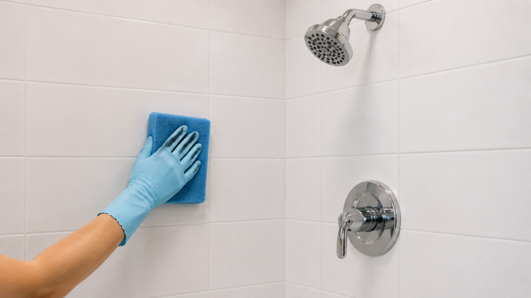 Gloved hand wipes a white tiled shower wall with a soft sponge beside a chrome showerhead and control.
