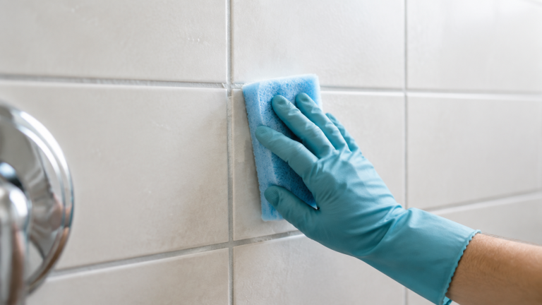 Gloved hand gently wipes white shower tile beside a grout joint with a soft blue sponge.