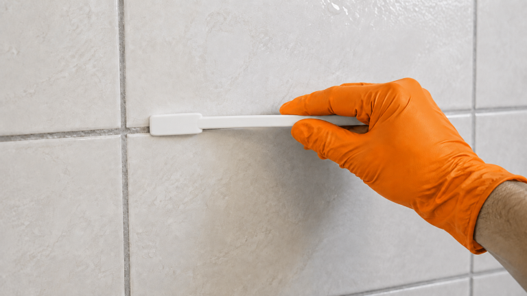 Orange-gloved hand holds a white cleaning brush against a grout joint between shower tiles.