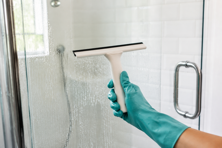 Teal-gloved hand uses a white squeegee to remove water from a glass shower door.